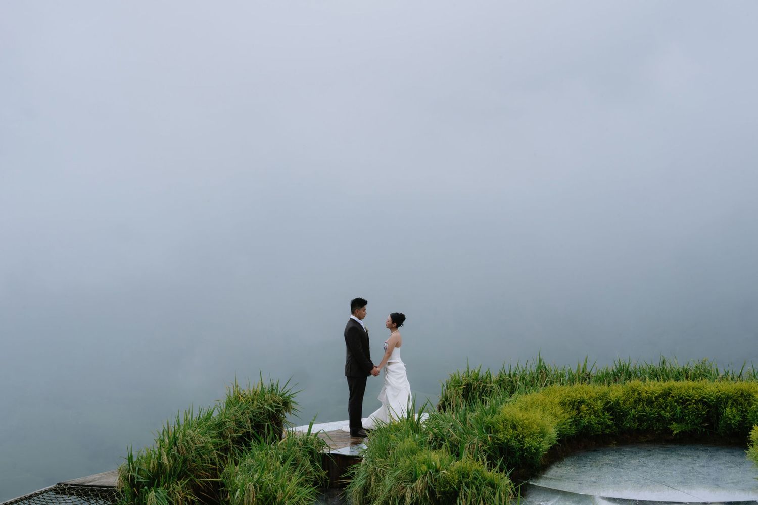 Bride and groom hold hands on misty overlook surrounded by lush greenery and fog.