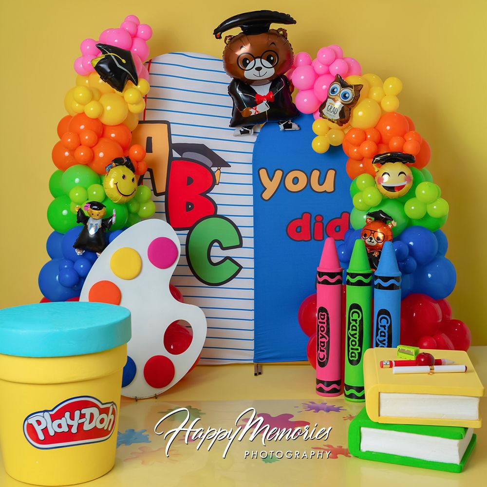 Colorful graduation party backdrop with balloons, ABC letters, crayons, Play-Doh, and a bear in cap and gown.