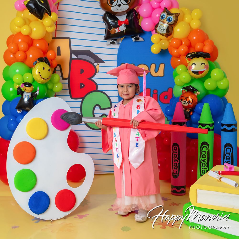 Child in pink graduation gown poses with oversized crayon and paint palette amid colorful balloon arch and school-themed backdrop.