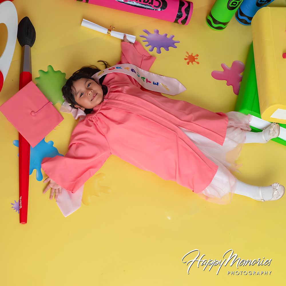 A girl in a pink dress lies on a yellow floor surrounded by oversized crayons and art supplies in a colorful photo.