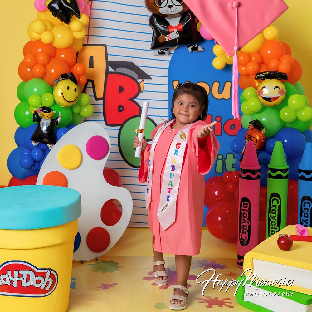 Young girl in pink graduation gown poses with diploma amid colorful balloon arch and Play-Doh, Crayola decorations.