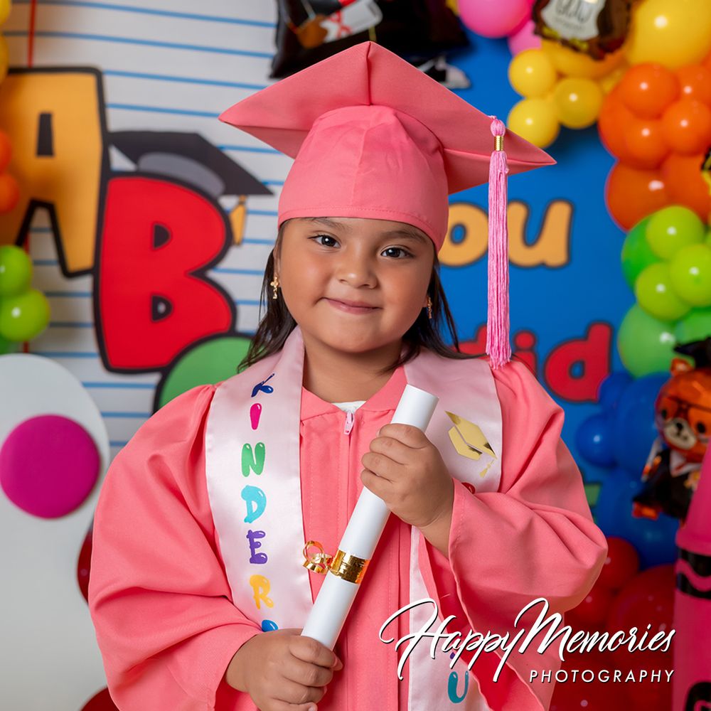 Young girl in pink kindergarten graduation cap and gown holding diploma scroll, smiling at colorful balloon backdrop.