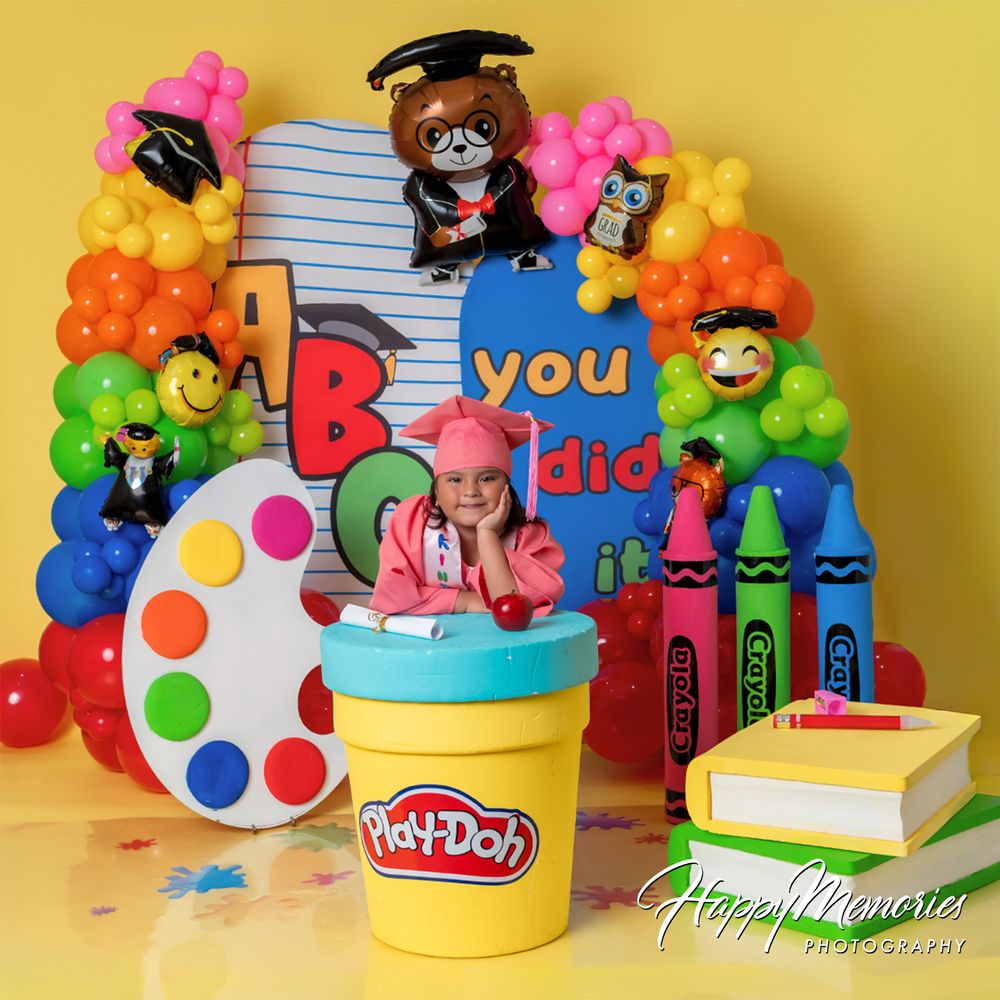 Child in pink hat sits in Play-Doh tub at colorful graduation party backdrop with balloons, crayons, and teddy bear.