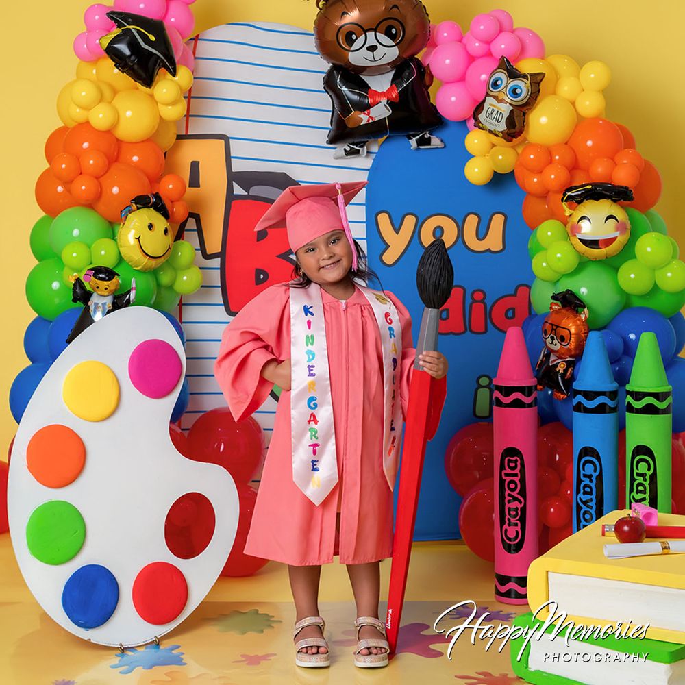 A young girl in a pink kindergarten graduation cap and gown poses with colorful balloons, crayons, and a paint palette backdrop.