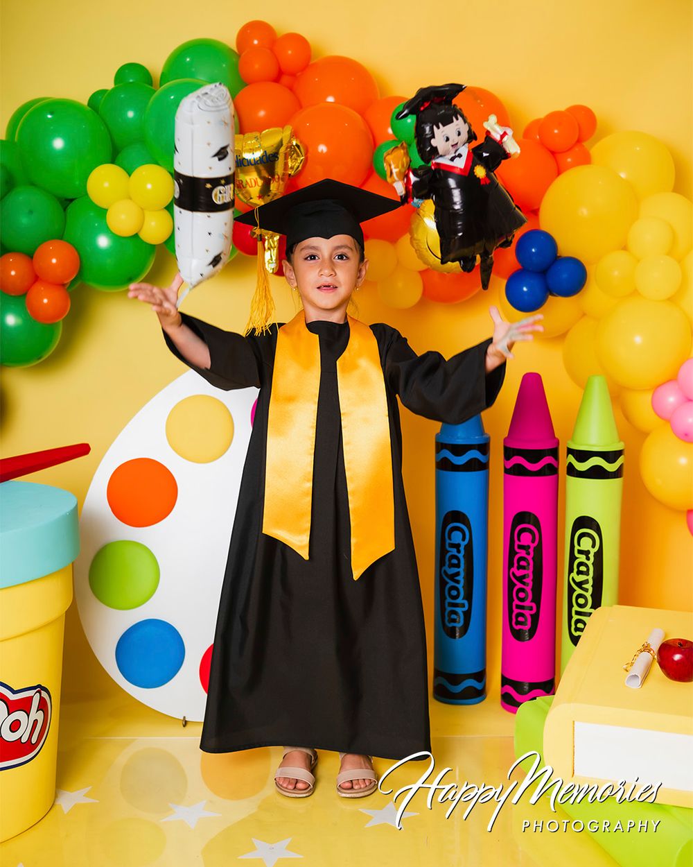 Colorful graduation themed photo setup with giant crayons, balloons, and cap and gown against yellow backdrop.