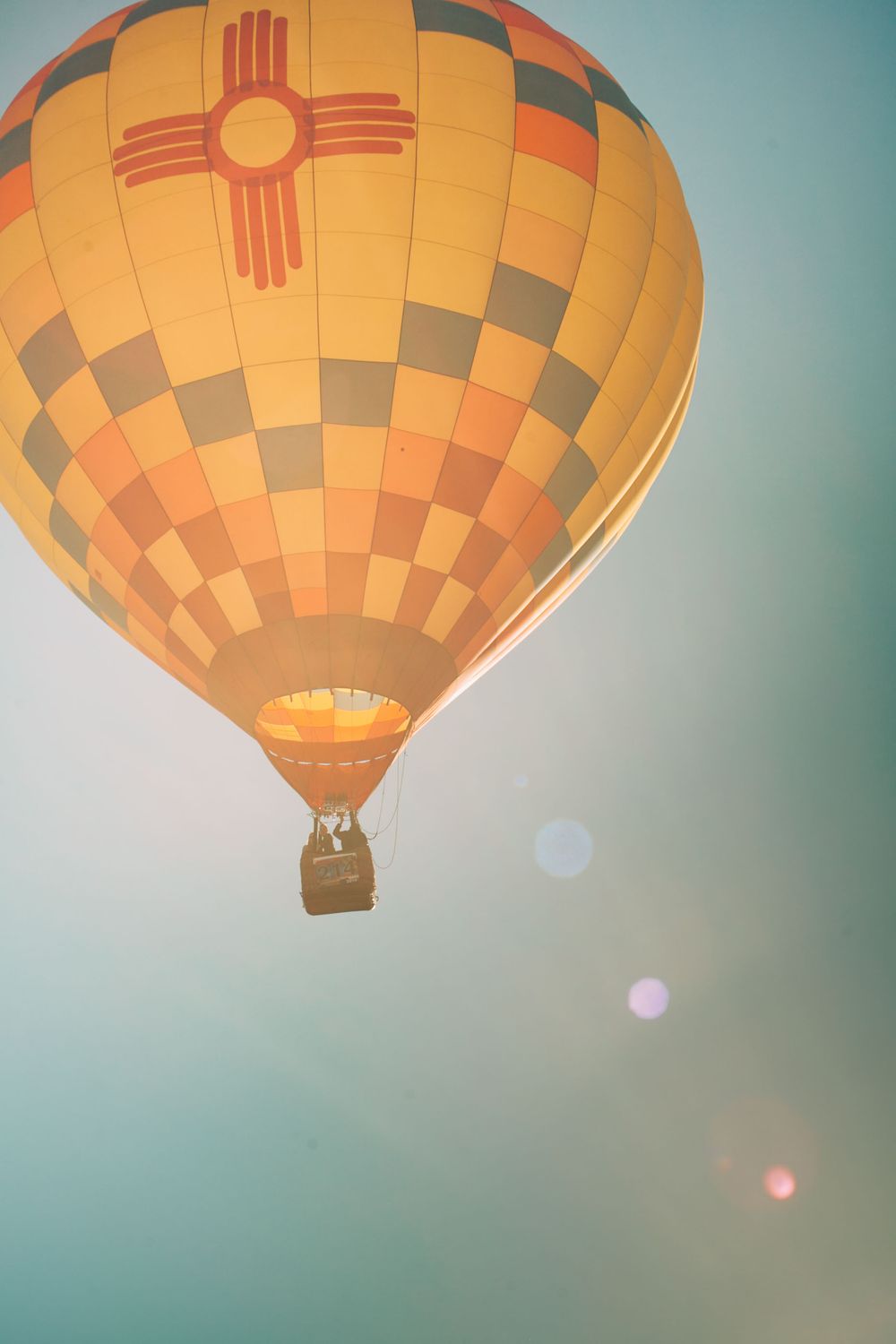 Hot air balloon with New Mexico Zia symbol floating against hazy sky during sunrise.