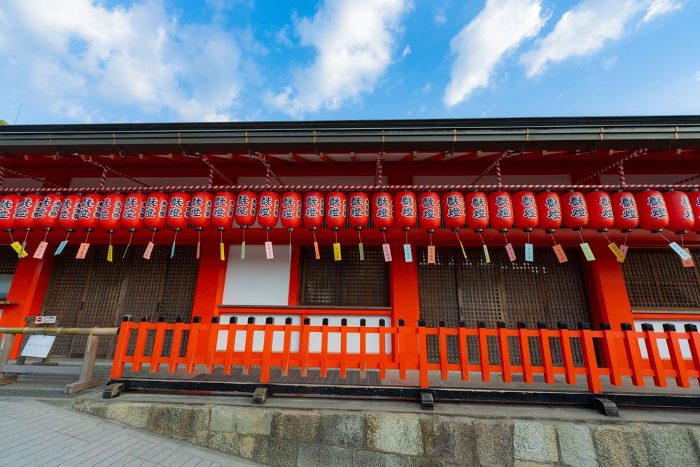 Row of bright orange torii gates and traditional Japanese architecture against blue sky with white clouds.