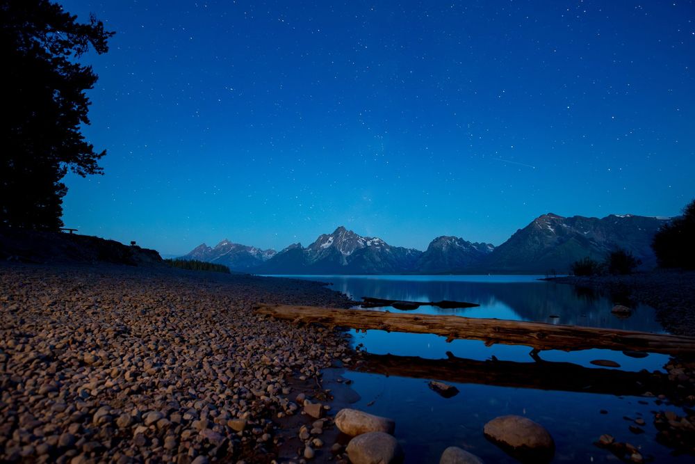 Mountain range silhouetted against deep blue sky with snow-capped peaks reflecting in calm lake waters.