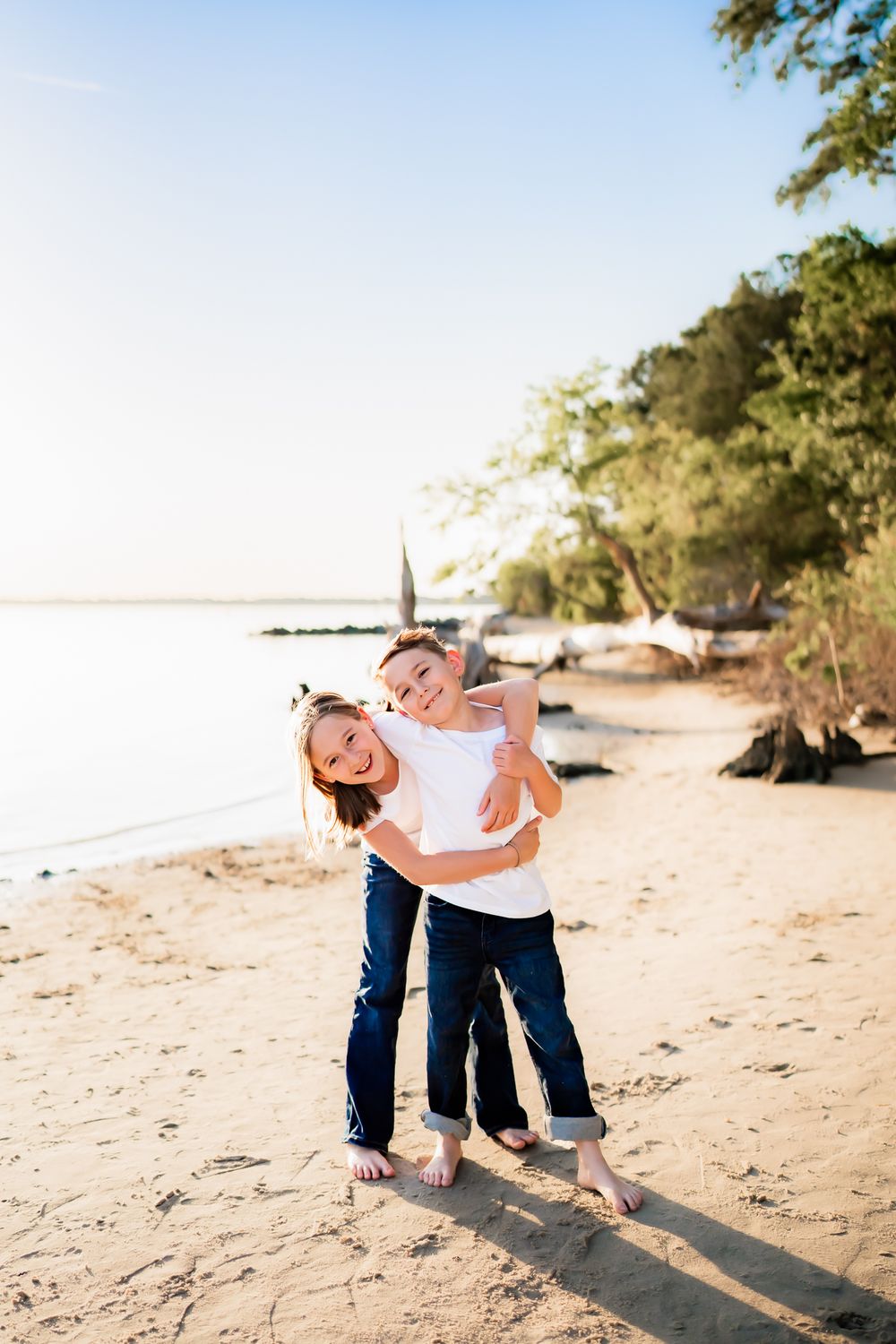 Couple in matching jeans embrace lovingly on a sunny beach with sand and trees in the background.