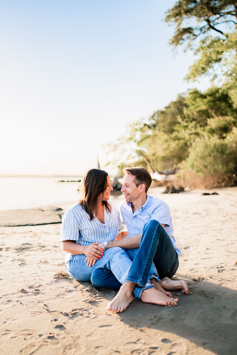 Couple sitting together on sandy beach at sunset sharing intimate moment near pine trees and shoreline.
