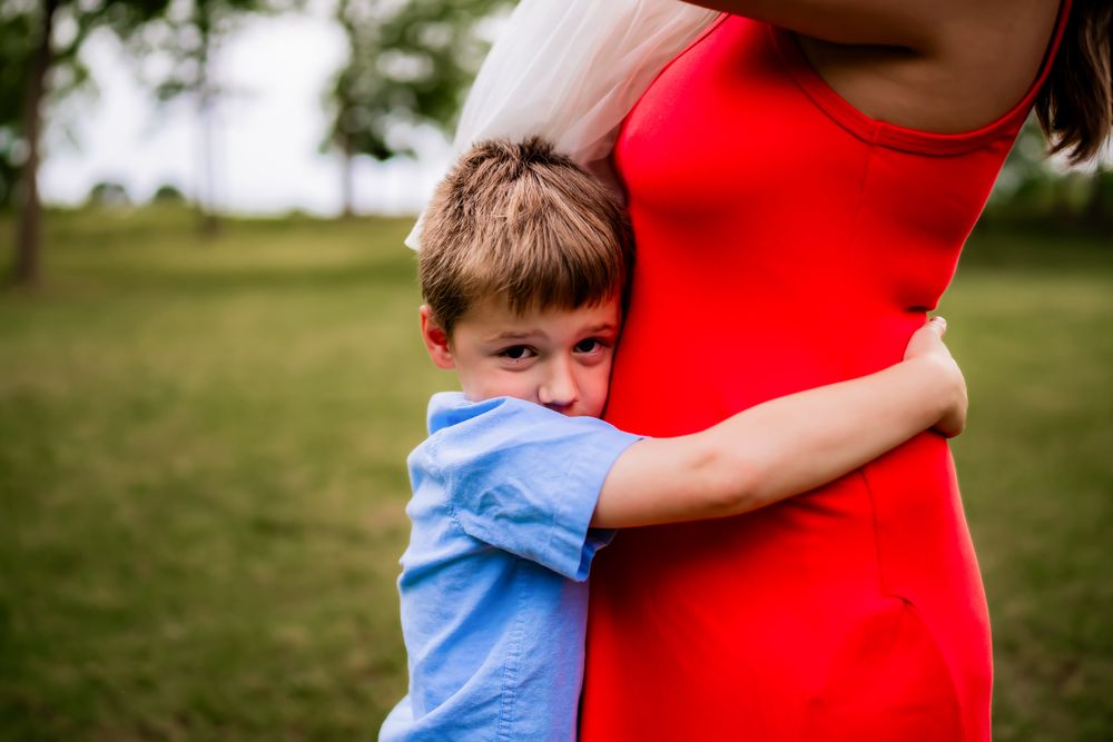Child in light blue shirt hugs someone wearing bright red in a sunlit outdoor setting.