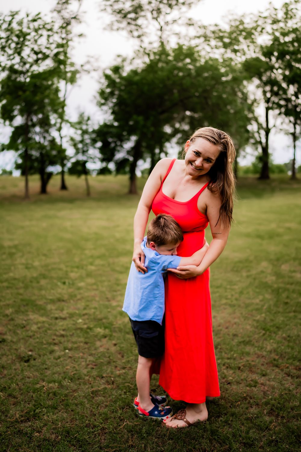 Mother in red dress shares tender outdoor moments with small child in blue shirt on grassy field.