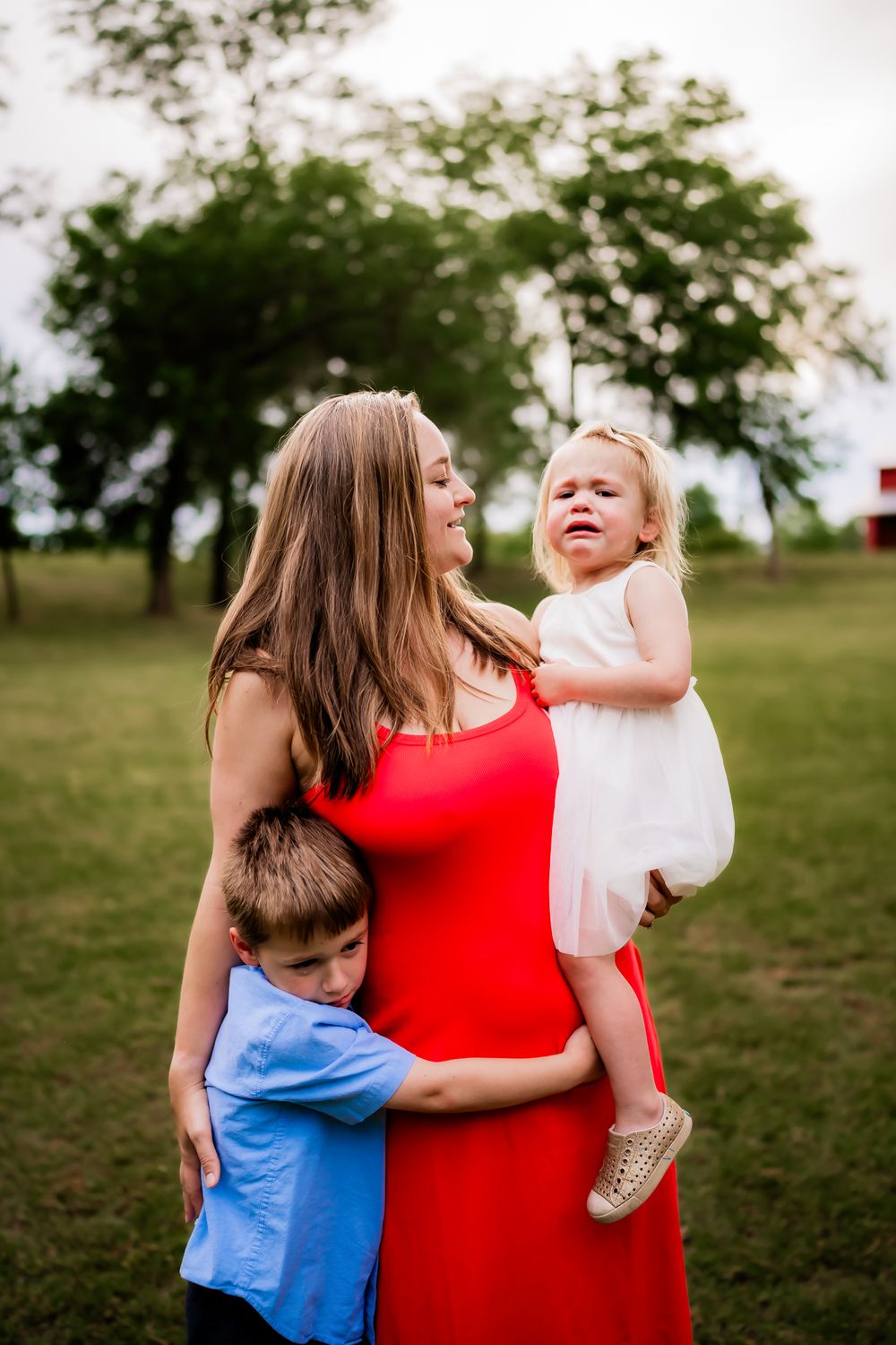A woman in a vibrant red dress shares tender outdoor moments with two young children in a grassy field.