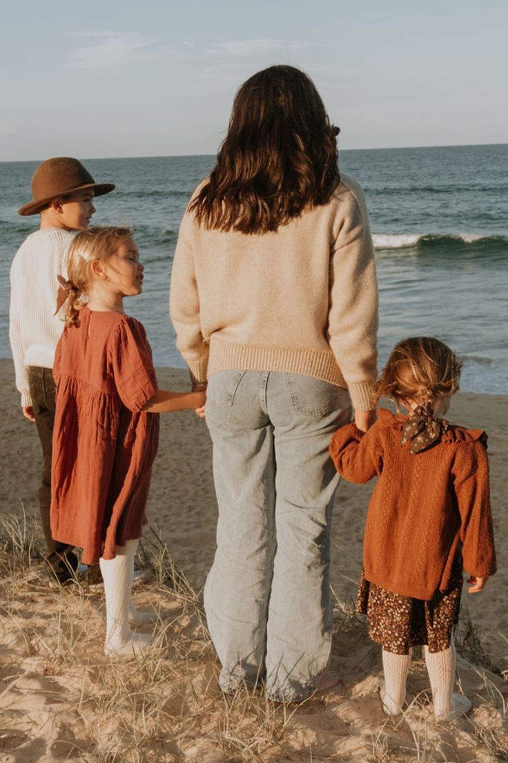 A family wearing earthy-toned clothing stands together on a beach looking out at the ocean waves.