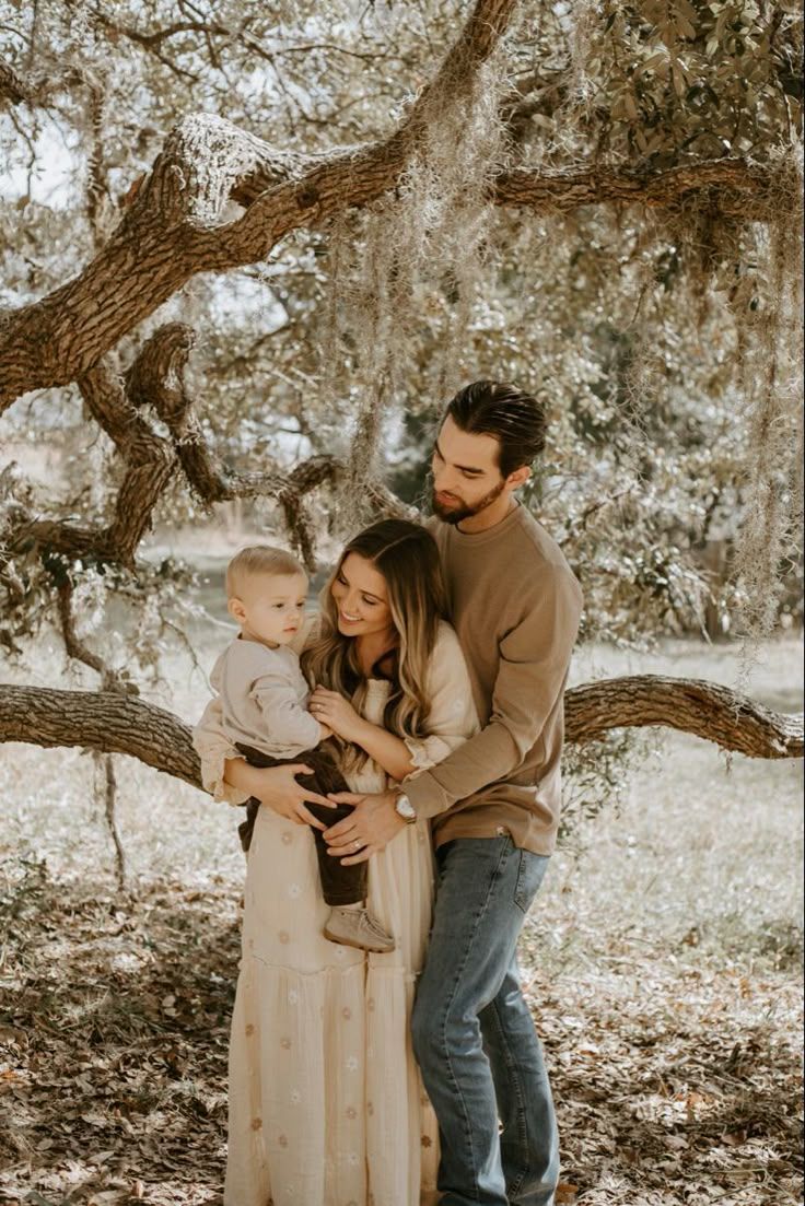 A small family shares a tender moment together under a large twisted tree branch in warm autumn lighting.