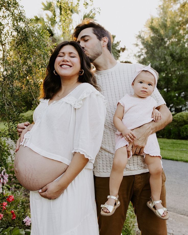 A family gathers outdoors on a sunny day, with a pregnant woman in white dress and a toddler wearing sandals.