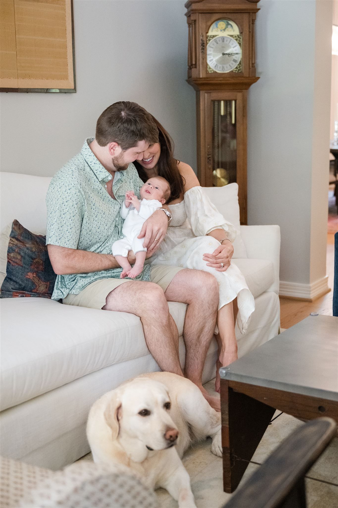 A person sits on a white couch playing with a baby while a yellow Labrador puppy rests nearby.