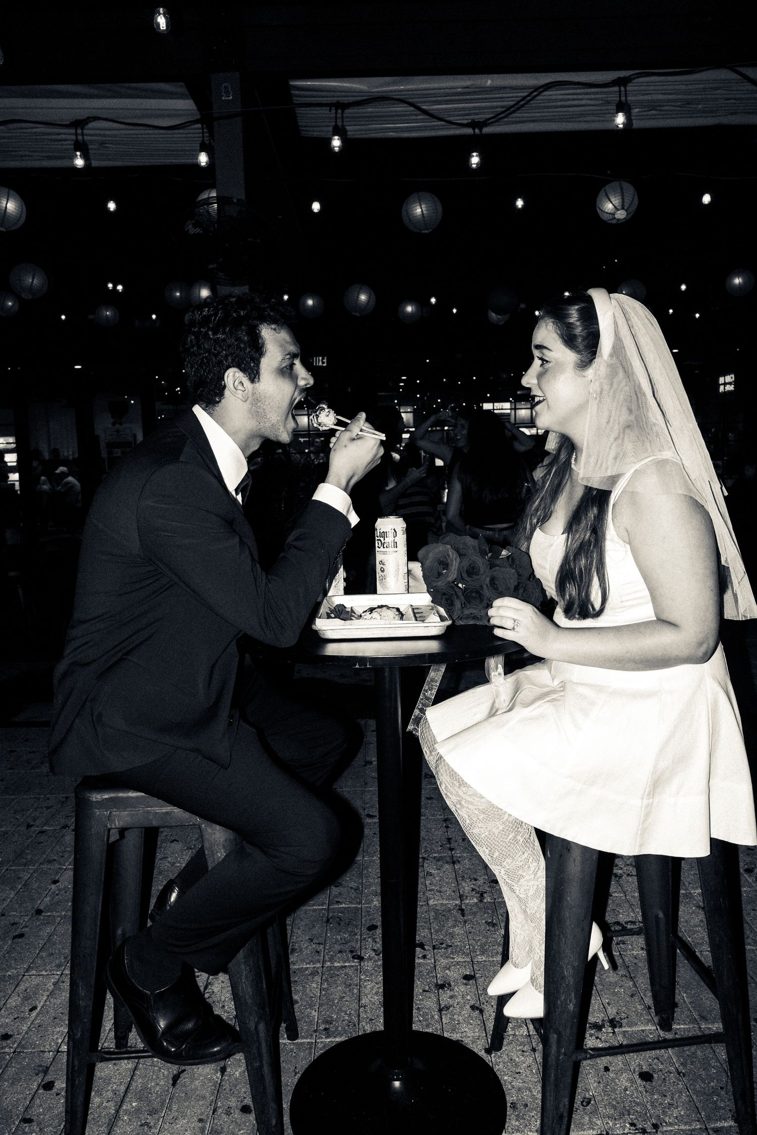 Wedding couple feeds each other cake playfully at the reception under string lights and red lanterns.