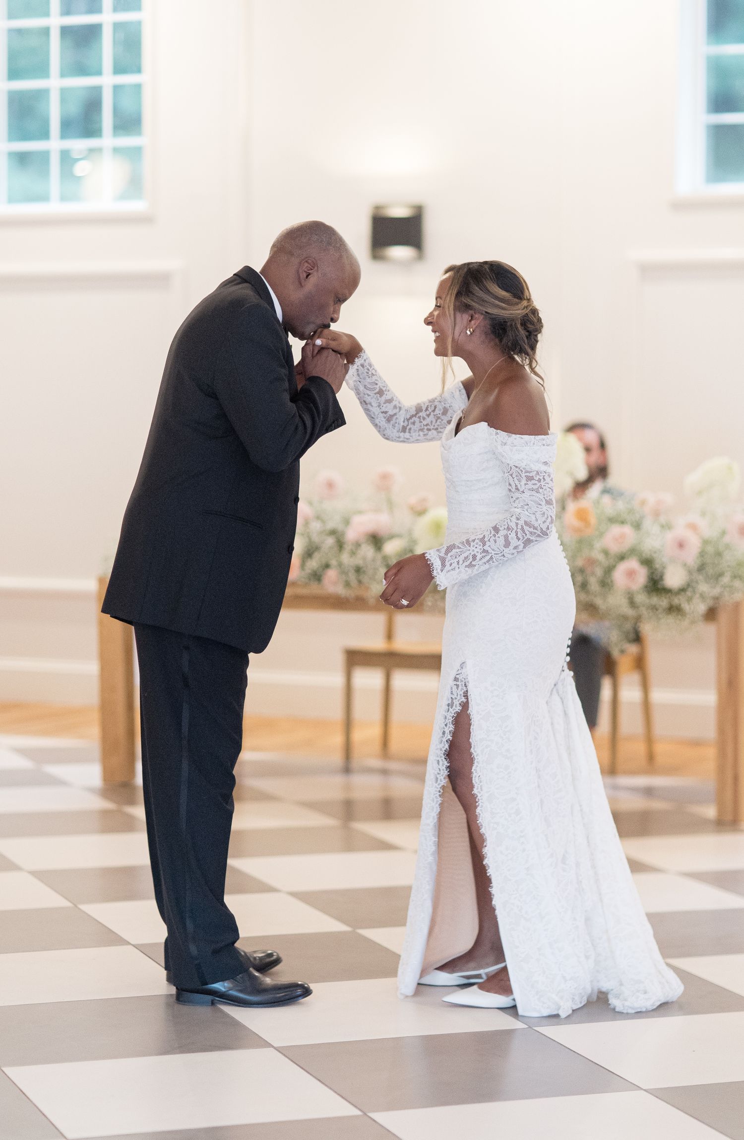 A couple in wedding attire shares a tender moment as the groom kneels before the bride in a light-filled room.