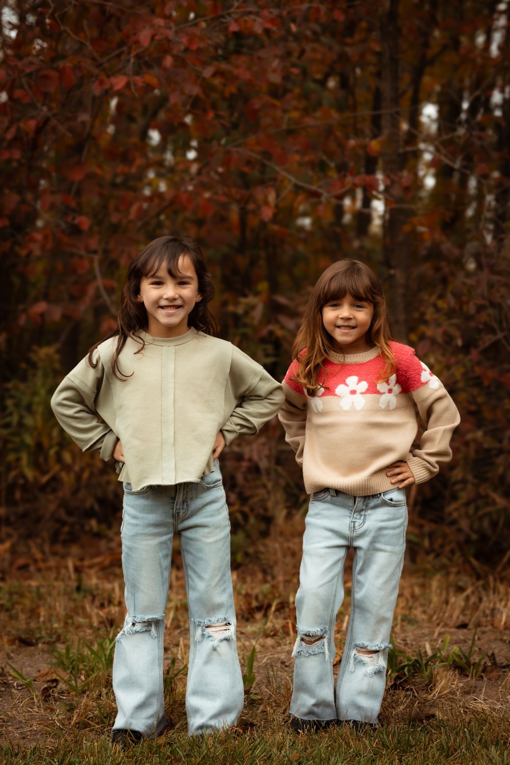 Two children wearing matching light wash jeans and winter sweaters pose outdoors on a grassy slope.