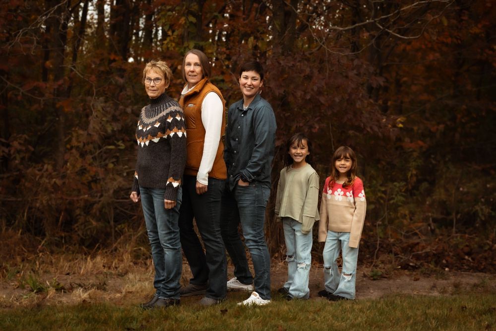 A group of five people stand together outdoors against a backdrop of autumn trees with brown leaves.
