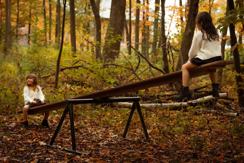 Two people balance on a seesaw in an autumn forest with fallen leaves and golden sunlight filtering through trees.
