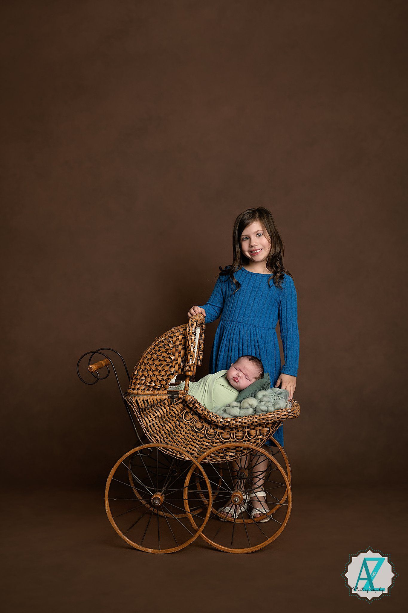 A young child in a blue sweater stands next to a vintage wicker baby carriage with a small infant inside against a brown backdrop.