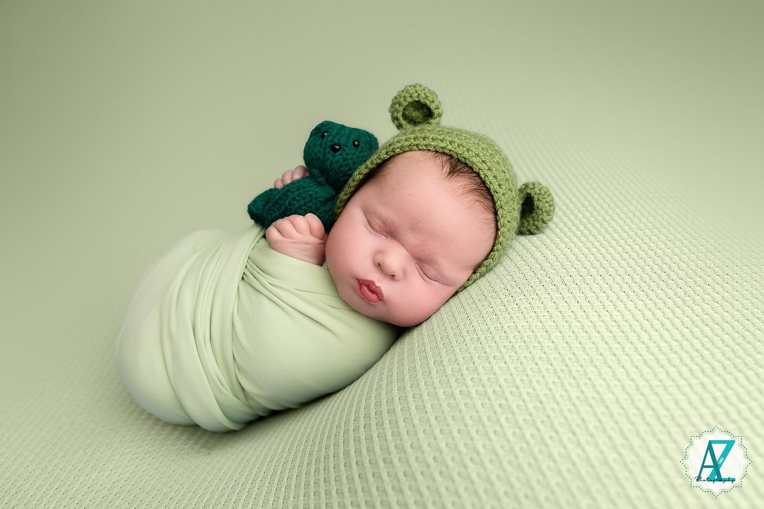 A sleeping newborn wearing a green knit hat is swaddled in soft white fabric against a textured white background.