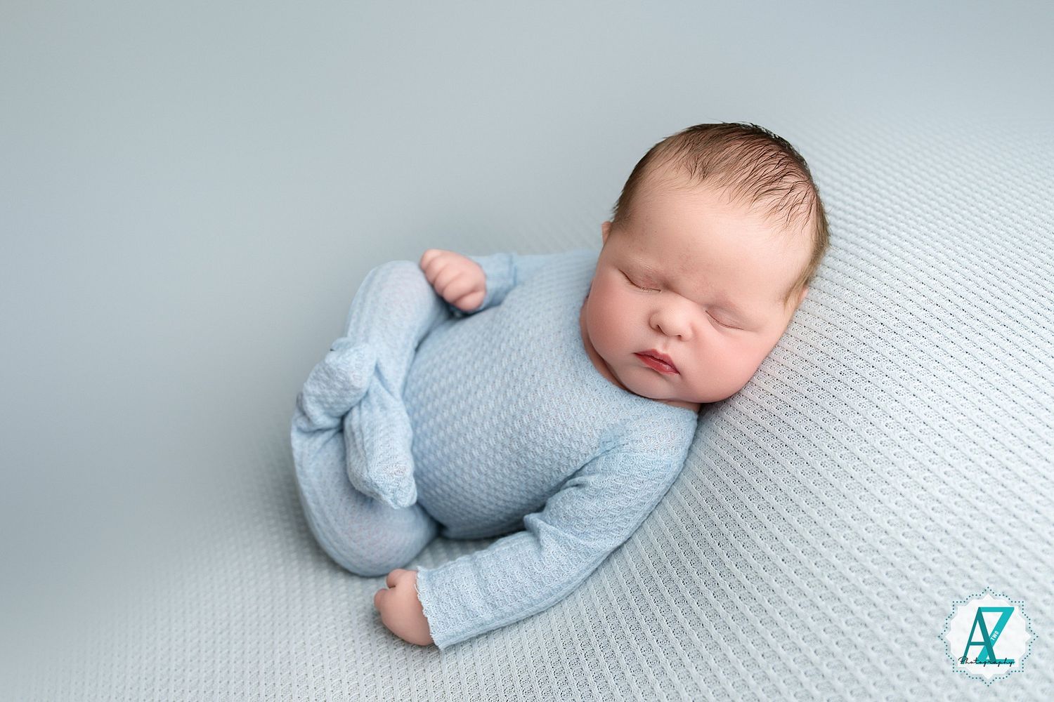 Sleeping baby wearing soft blue outfit lies peacefully on textured white fabric in studio lighting.