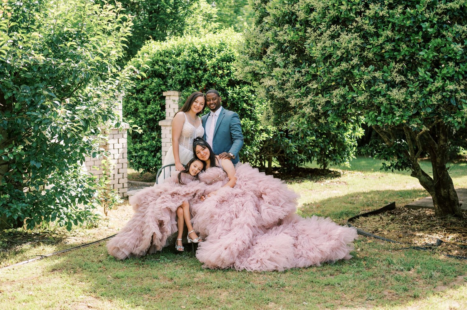 A couple in formal attire poses in a garden setting with a flowing pink tulle gown spread across the grass.