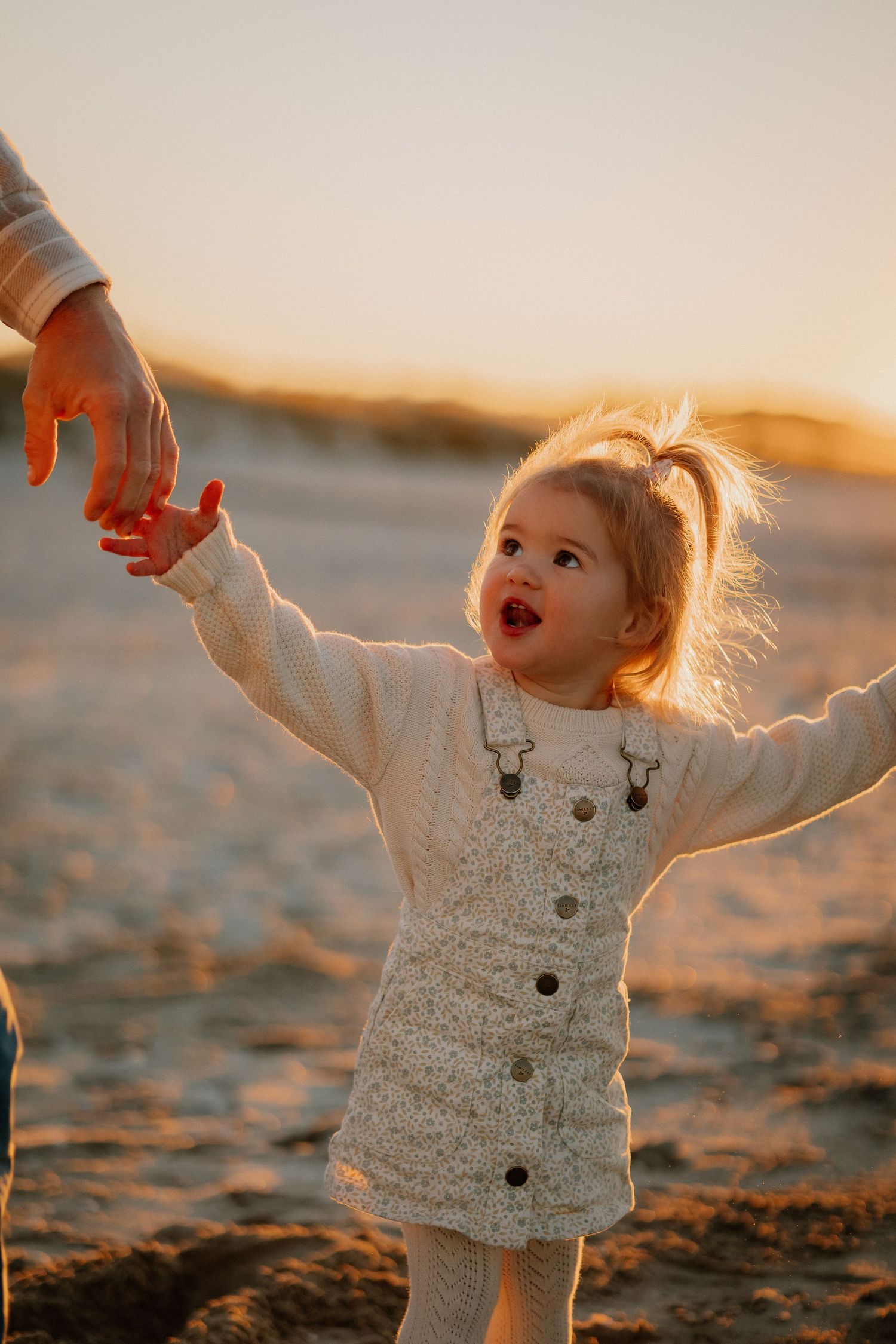 A toddler in a white sweater playing joyfully on a sandy beach at sunset as their hair glows in golden light.