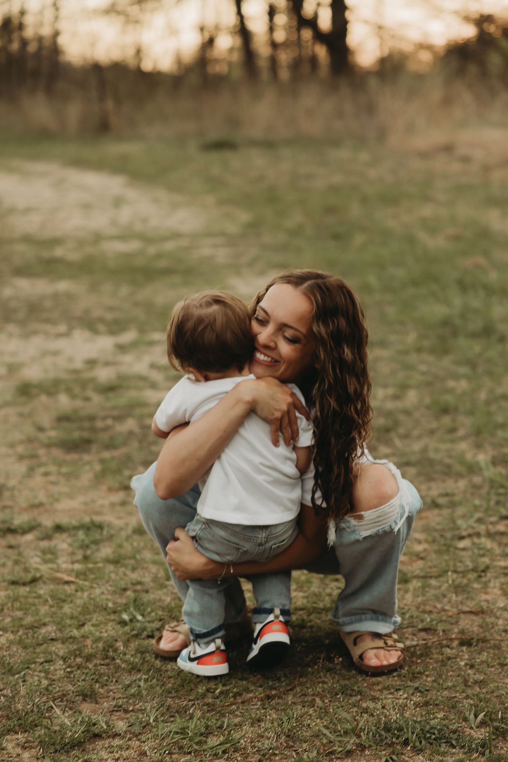 Mother and child share tender embrace while sitting together on grass during sunset photoshoot.