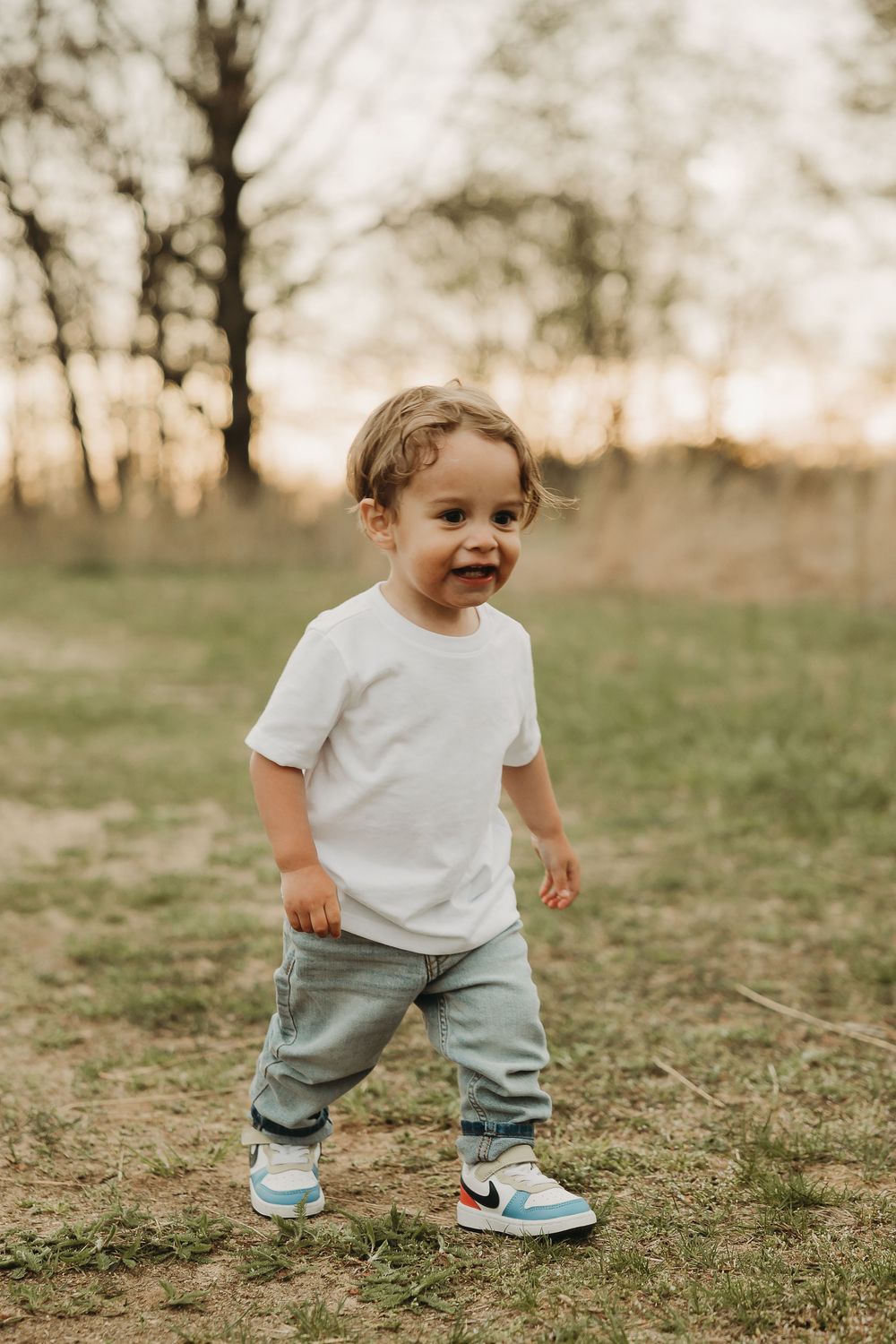 Small child in white shirt and blue pants walks through grassy field at sunset wearing colorful sneakers.