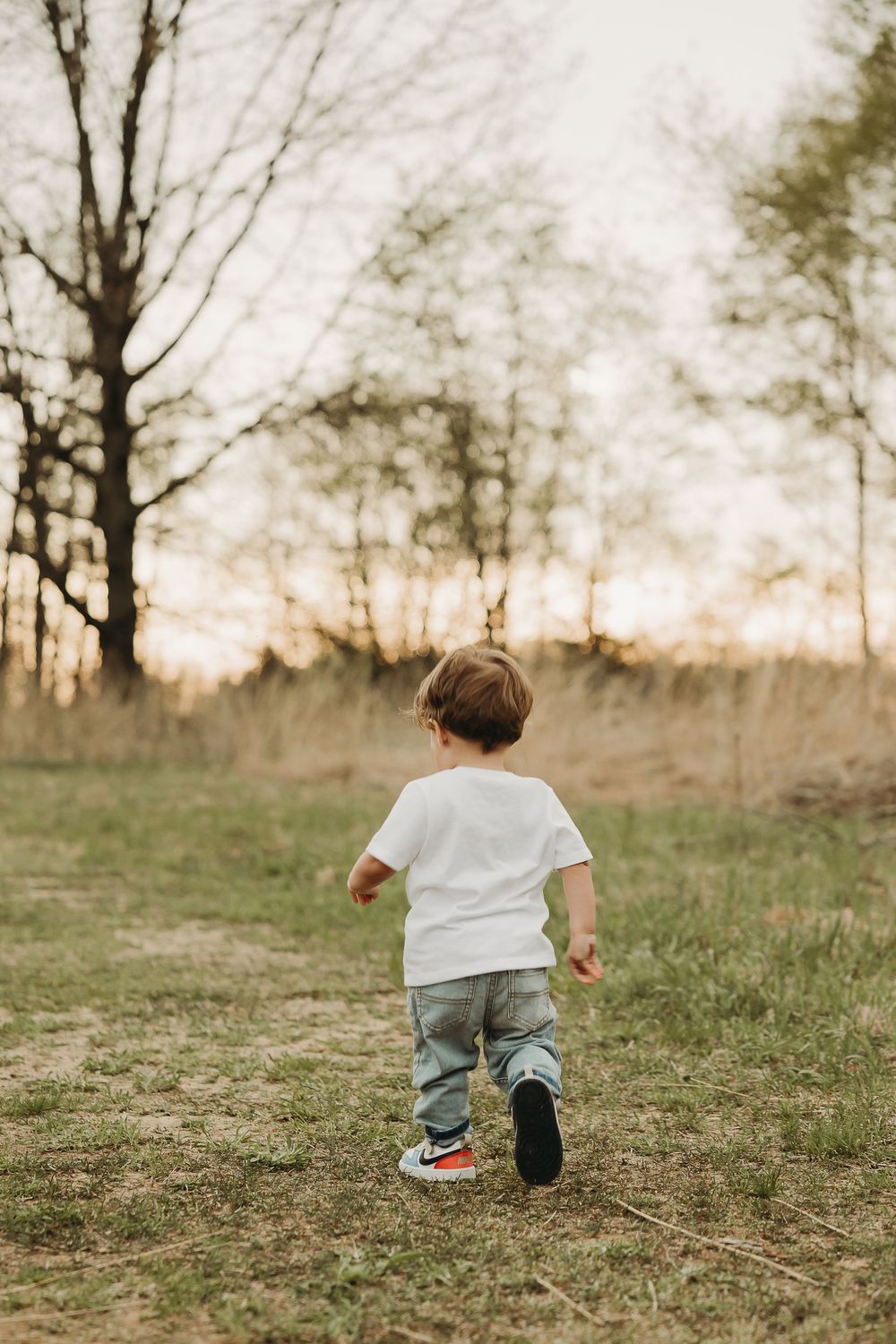 Small child in white t-shirt and jeans running through a grassy field at sunset with bare trees in background.