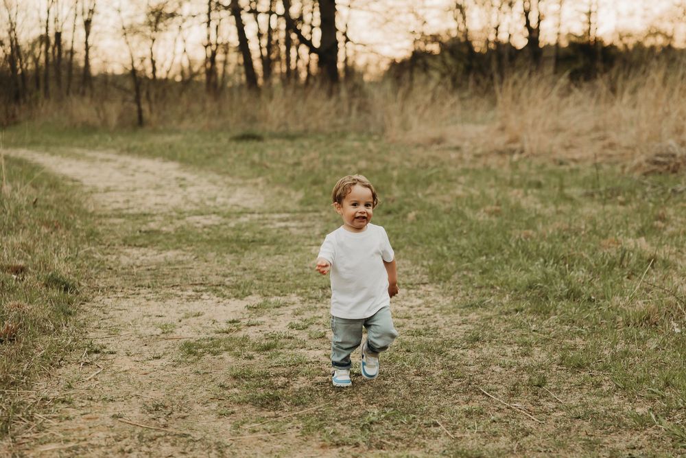 A toddler in jeans and a white shirt walking along a dirt path in a grassy field at sunset.