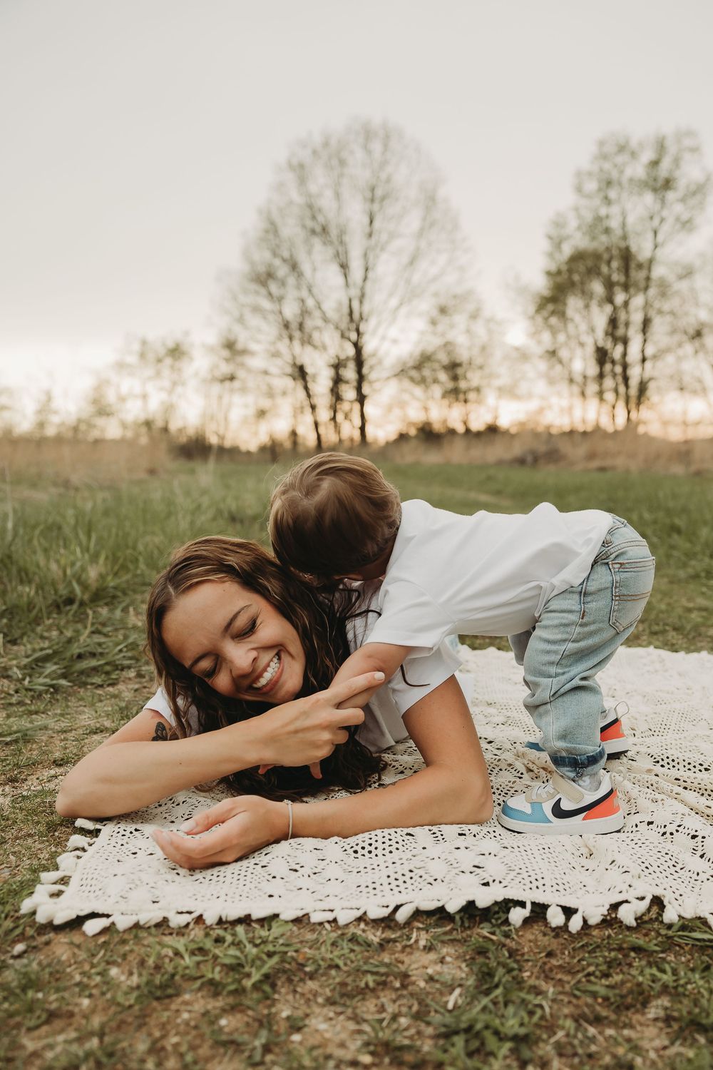 A playful moment captured on a white lace blanket outdoors with trees silhouetted against an overcast sky in the background.