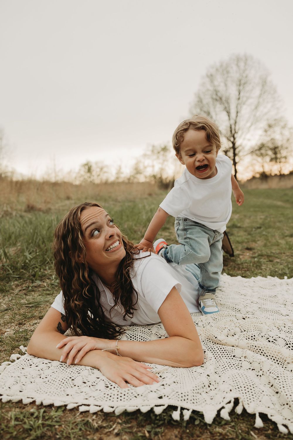 Two people share a joyful moment together on a white blanket in a grassy field at sunset.