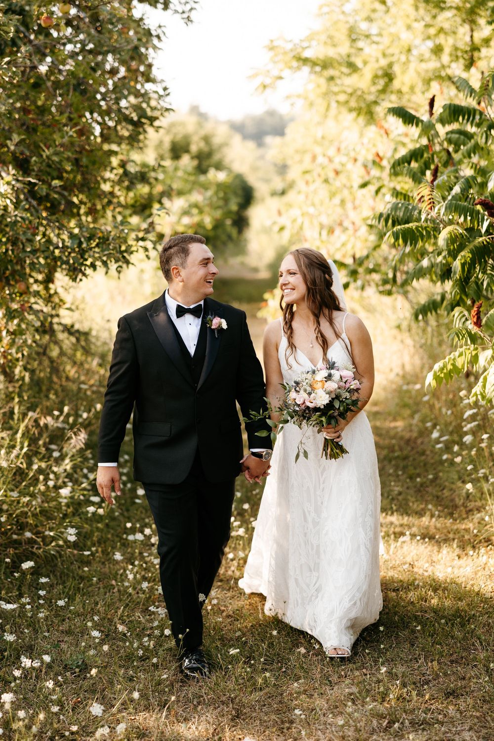 A couple walks together on a sunlit garden path, the bride in white lace carrying a bouquet while sharing a joyful moment.