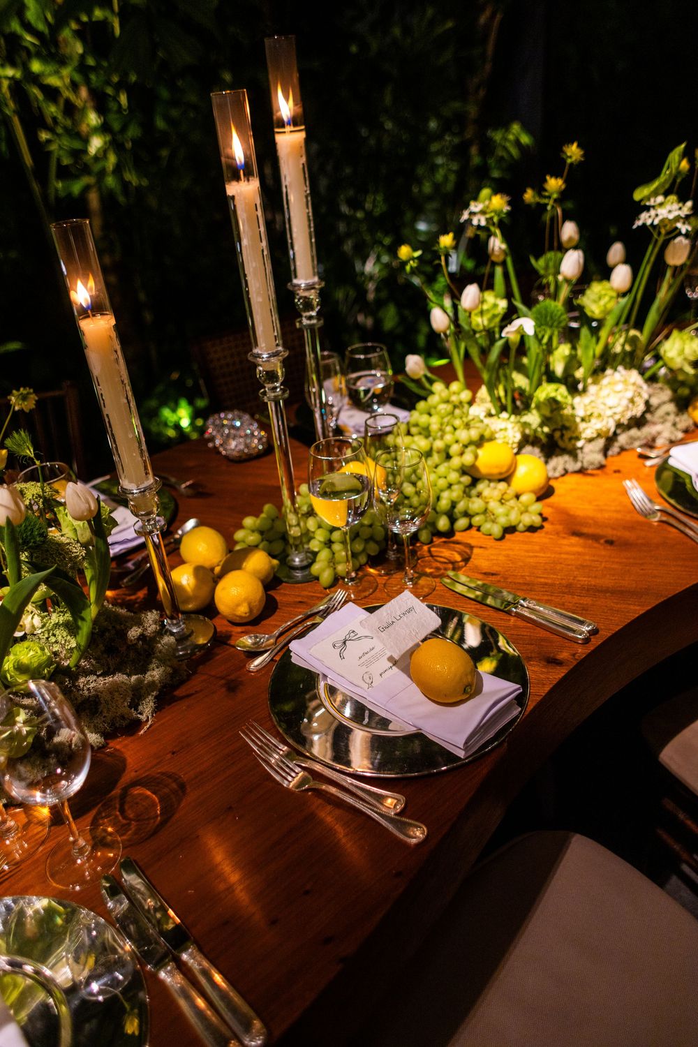 Elegant table setting with candles and citrus fruits on dark wooden table decorated with fresh greenery at night.