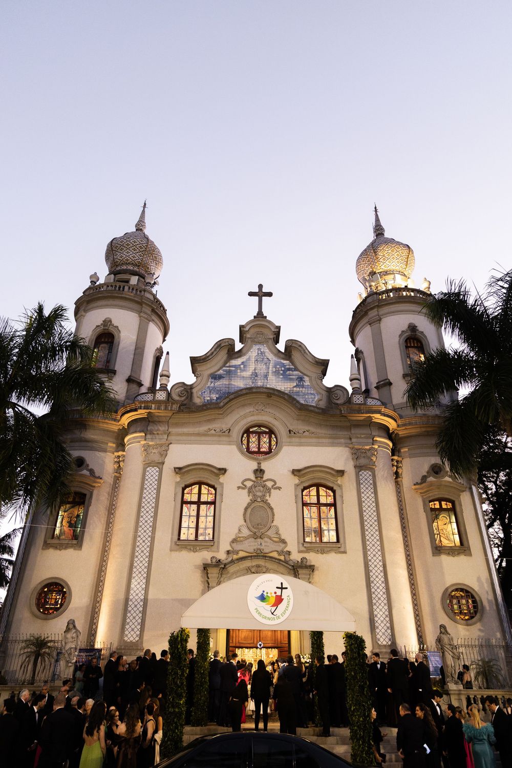 White Spanish colonial church facade illuminated at dusk with palm trees and gathering crowd outside.