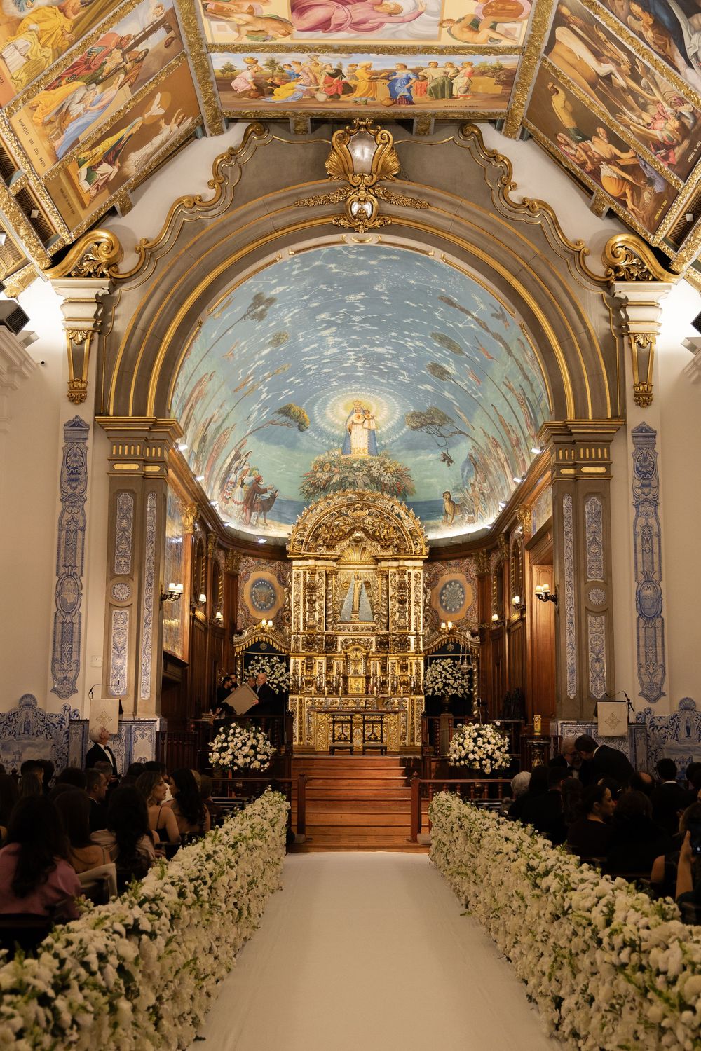 Ornate church interior with white floral-lined aisle leading to a grand golden altar under a painted baroque ceiling.