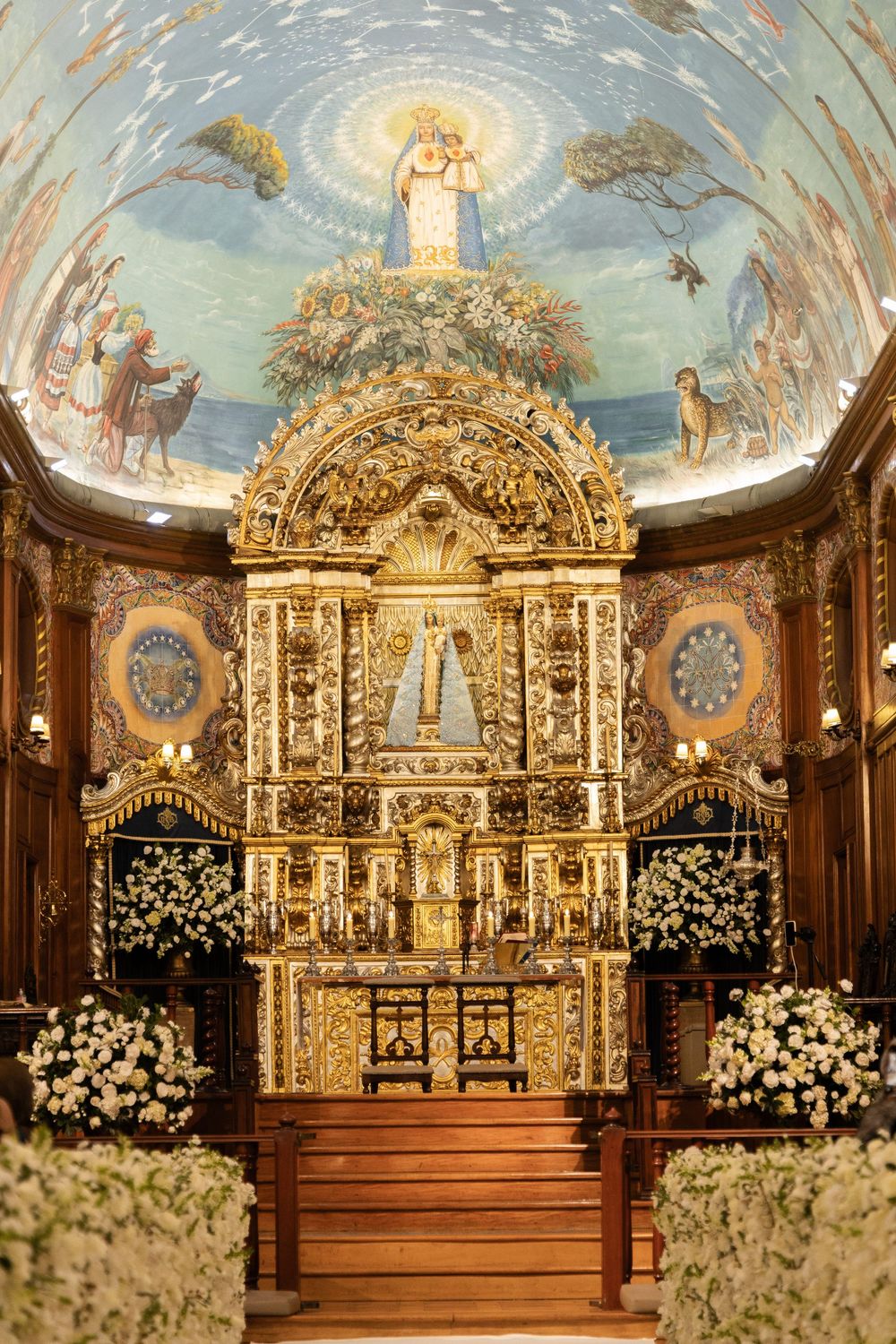 Ornate golden baroque church altar with painted ceiling murals and white floral decorations in an elegant sanctuary setting.