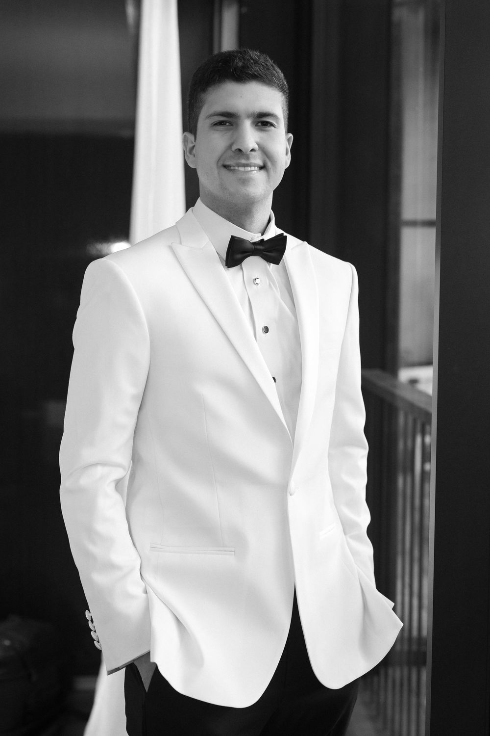 Black and white portrait of someone in a classic white tuxedo jacket with black bow tie standing outdoors.