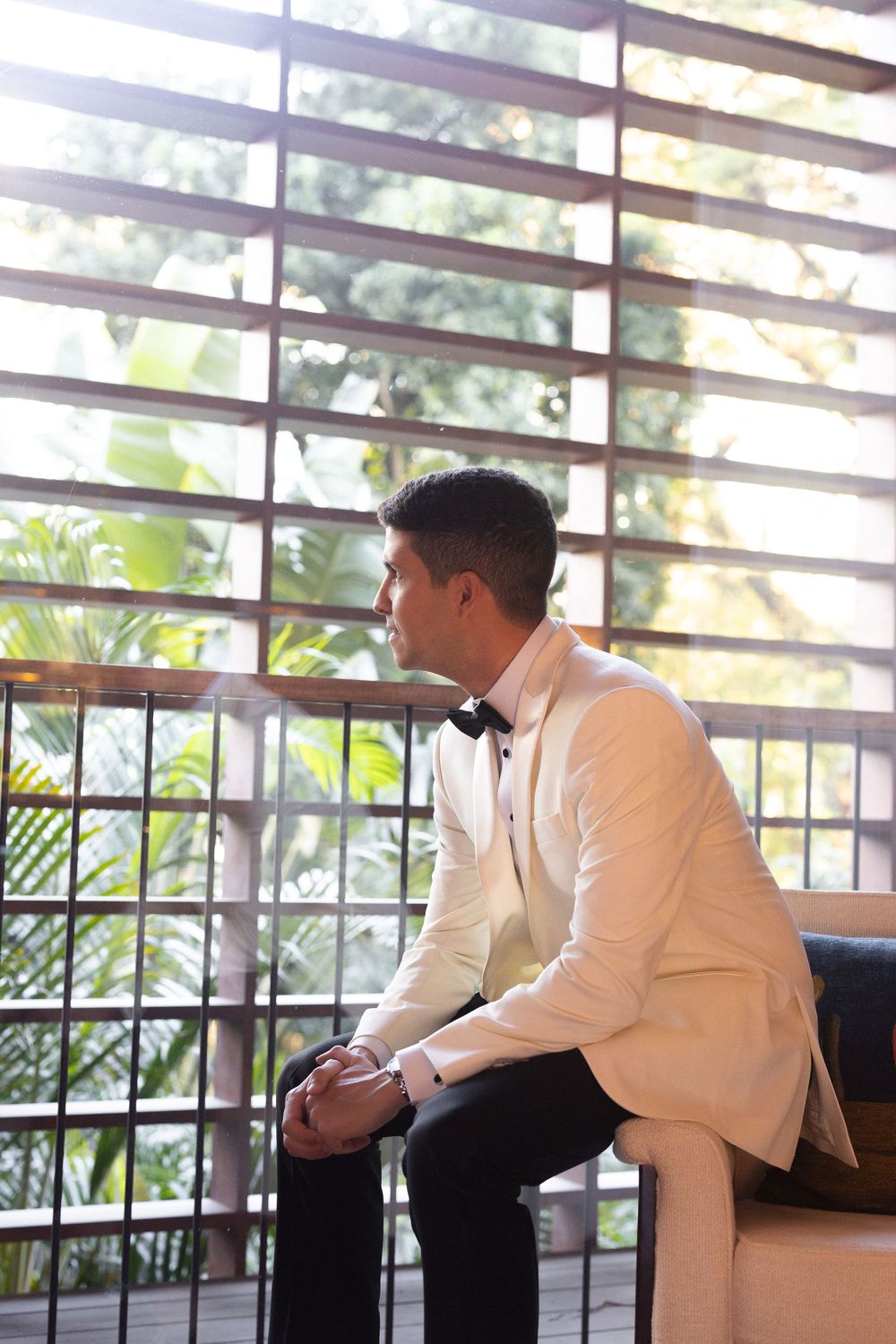 A person in a white shirt sits thoughtfully on a bench against a window with slatted blinds and tropical foliage outside.