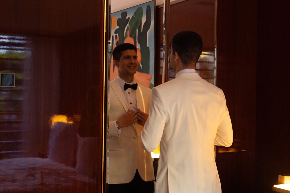 A person in a white jacket holding a glass of wine stands in front of a dimly lit wooden wall.