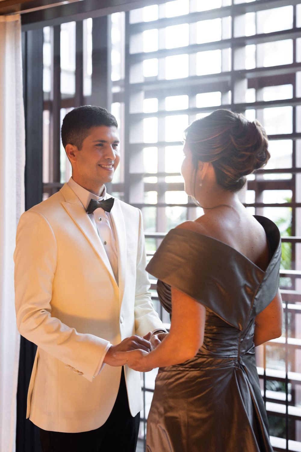A couple in formal attire with white tuxedo and brown dress dancing together in a bright sunlit room.