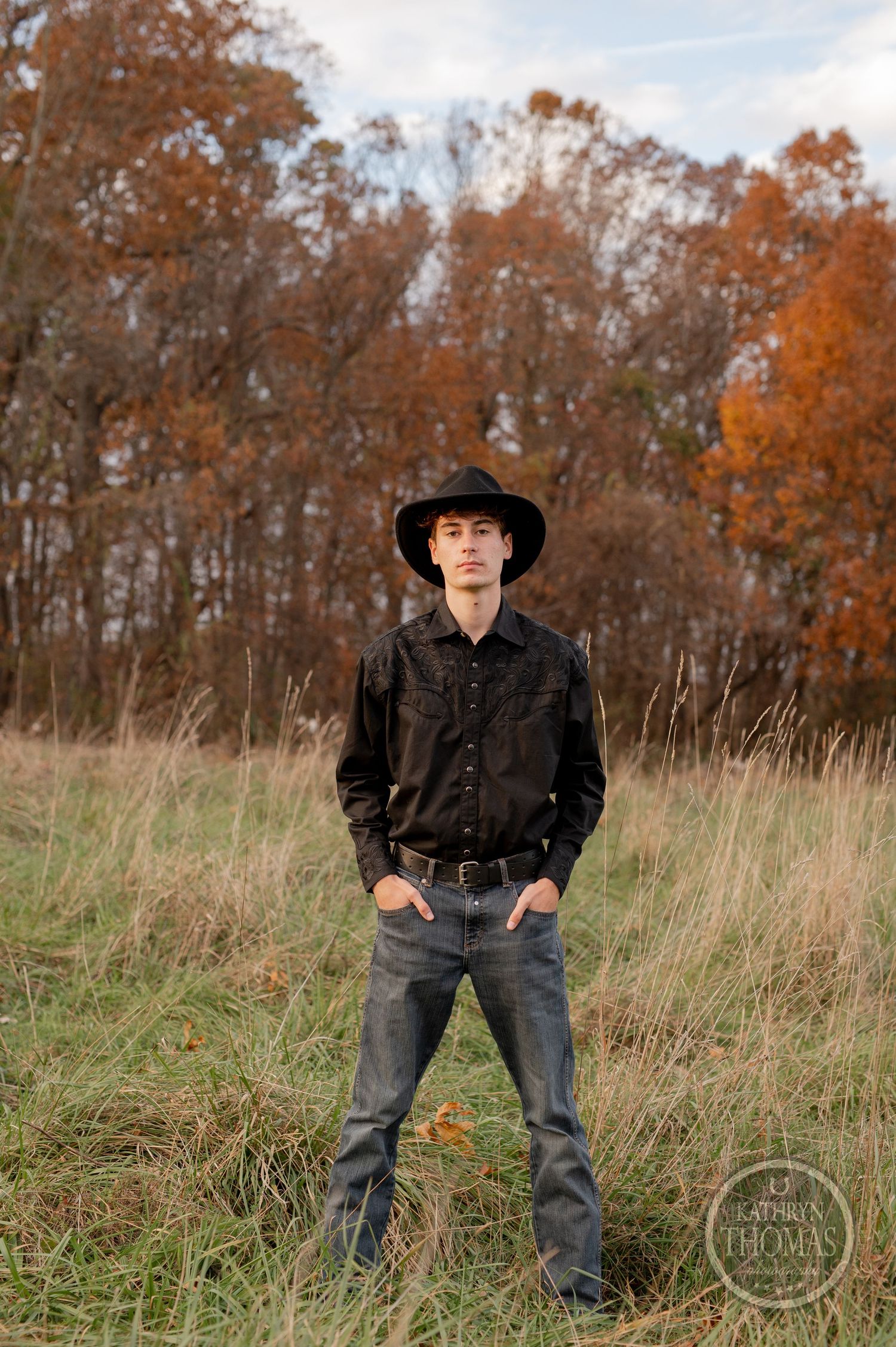 Standing portrait in western attire against a backdrop of autumn trees and tall grass.