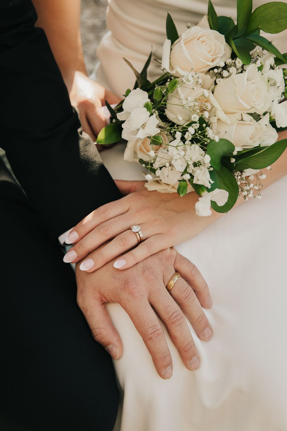 Wedding bouquet of cream roses and white flowers next to clasped hands displaying matching wedding rings and manicured nails.