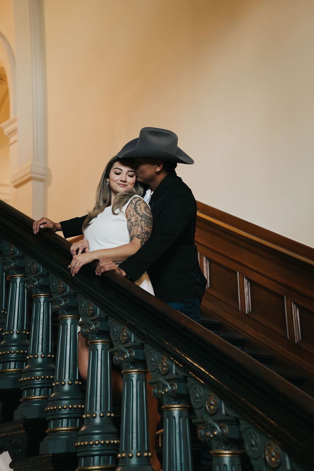 Romantic couple in western attire share intimate moments on an ornate vintage staircase with dark wood details.