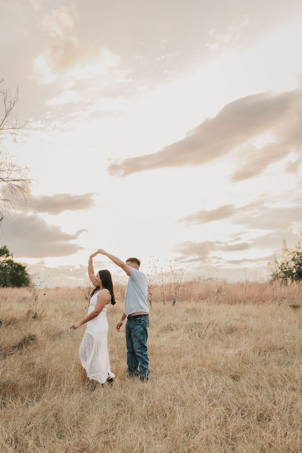 A couple dancing in a golden field at sunset with dreamy clouds overhead.